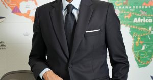 Man in a dark suit and tie standing in front of a colorful world map